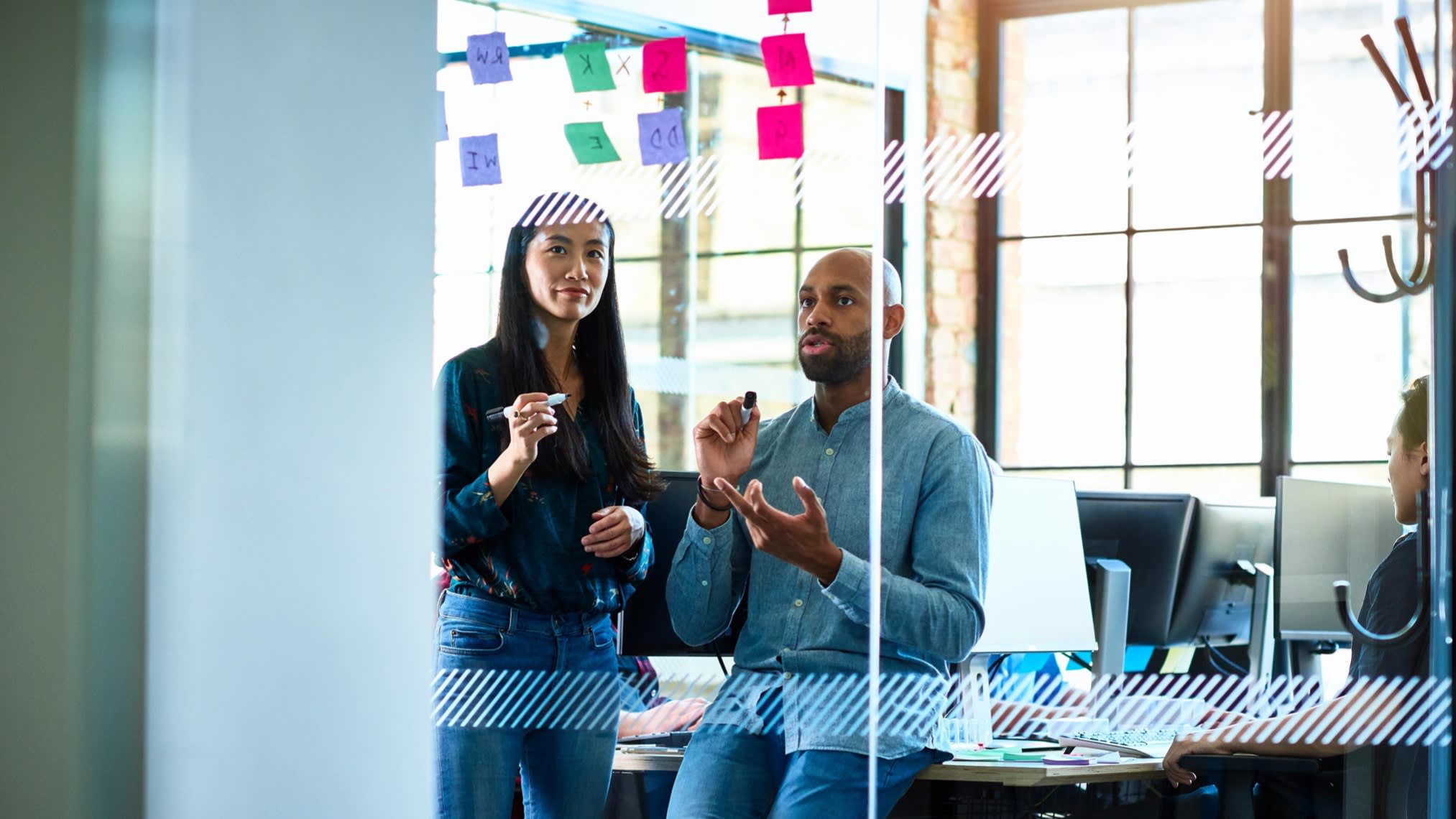 Asian woman and African American colleague talking and looking at post it notes, stuck to glass partition